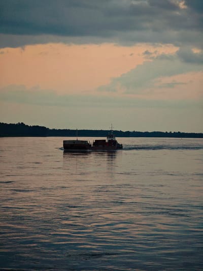 A barge is traveling on a wide river, moving from left to right in the frame. The barge appears to be hauling cargo. The water has a rippling surface, reflecting the muted colors of the sky. In the background, a dark line of trees forms the horizon. The sky above is filled with clouds, with shades of gray and pale pink. The scene suggests either sunrise or sunset. The location is identified as the city of Kazachinskoe, Russia.