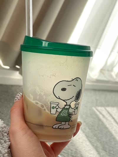 An indoor scene depicts a person holding a clear plastic reusable tumbler with a green lid and a transparent straw. The tumbler contains a light brown, milky beverage with ice cubes, and condensation is visible on the interior upper surface. A black outline cartoon of Snoopy, wearing a green apron labeled "JOE KIND" and holding two cups with green logos, is printed on the tumbler. A human hand, with a thumb displaying a white base with black polka-dot nail polish, grips the lower portion of the cup. The background is out of focus but shows light-colored curtains or blinds, with strong natural light entering from the right, illuminating a soft, textured grey fabric surface below. The precise location in Wanneperveen, Netherlands, is not discernible from the visual evidence.