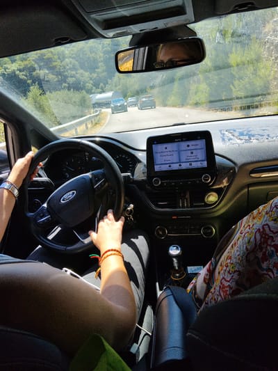 The image shows the interior of a Ford vehicle in motion, viewed from the passenger seat. The driver's hands are on the steering wheel. The car is traveling on a road that appears to be in a rural or mountainous area. In the background, there are trees and a clear sky, suggesting it is daytime. A truck is visible in front of the car. The dashboard displays a touchscreen infotainment system showing music playback and navigation options. The exterior environment is consistent with the hills surrounding Mattinata, Italy, a coastal town known for its scenic landscapes.