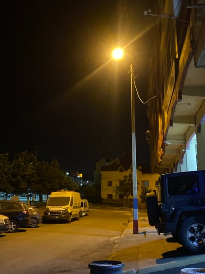 The image depicts a street scene at night in El Hajeb, Morocco, illuminated by a bright streetlamp. In the foreground, a dark-colored Jeep is parked on the right, with its spare tire visible. To the left, a white ambulance is parked in front of other vehicles. The ambulance has text on its front, which appears to be in Arabic script and reads "Ambulance" in reverse. Behind the ambulance, several trees are visible. Further back, buildings with distinctive architecture, including arched doorways and windows, line the street. The overall environment suggests a residential or commercial area within the city. There are no people visible in the frame.