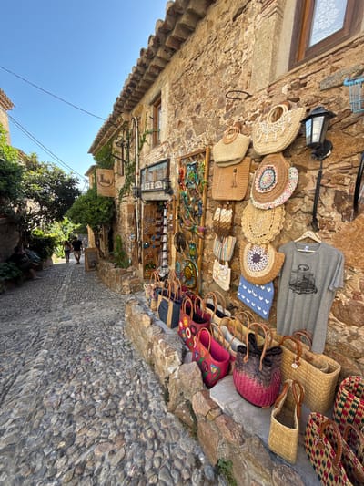 This image depicts a narrow, cobblestone street in Tossa de Mar, Spain, lined with stone buildings. The scene is an outdoor market or shop display. Various handmade items, primarily woven bags and hats, are displayed hanging on a stone wall and on shelves. A grey t-shirt with a bicycle graphic is also visible, hanging from a hook. Some of the bags are intricately patterned with colorful threads, while others are made of natural straw. There are also some embroidered shoes or slippers with floral designs. In the background, a few people are walking down the street. The sky is a clear, bright blue, suggesting a sunny day. The architecture and cobblestone street indicate a historic European town. A sign with text, partially visible, reads "CHEVEURES." The overall impression is one of a charming, artisanal marketplace bathed in sunlight.