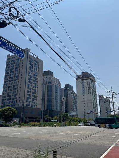 The image is taken from a street-level perspective at an intersection in Pyeongtaek-si, South Korea. The foreground shows a paved road with white lane markings. In the middle ground, there is a green bus turning right, a white car and a truck stopped at the intersection, and various buildings, including apartment complexes and a hotel, as well as trees and vegetation. Overhead, a complex network of electrical wires is visible against a clear blue sky. A blue street sign with Korean and English text reads "평택항로 Pyeongtaekhang-ro 209". Traffic lights and a speed limit sign (50) are also present. The overall scene depicts a typical urban streetscape on a sunny day.