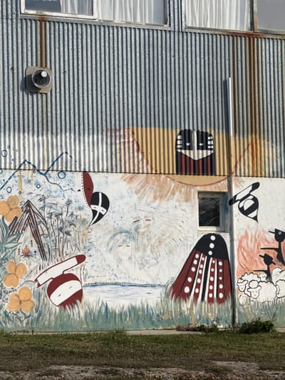 A section of an exterior building wall is depicted in Tolhuin, Argentina, illuminated by direct sunlight. The upper portion of the wall consists of vertical, corrugated galvanized metal siding, exhibiting areas of rust and discoloration. Two windows with white curtains are visible at the very top. A circular, louvered metal ventilation fan is affixed to the corrugated siding on the upper left. A rectangular segment of the corrugated metal in the upper right features a painted stylized face or mask, rendered in black, white, and red.
The lower two-thirds of the wall are covered by a multi-panel mural painted over a light base coat, showing signs of wear and paint flaking. The mural depicts various scenes and stylized figures. On the left, a landscape features blue lines suggesting mountains, yellow-orange flowers, and green foliage. A triangular, dark-brown structure resembling a dwelling stands among sparse trees, with small, stick-figure human forms nearby, one appearing to tend a small fire. A red and white abstract shape is present in the lower-left. Above the center-left, a stylized black and white mask-like shape with a red outline is painted.
The central part of the mural portrays several figures with long, flowing white hair and red facial markings, set above a blue body of water. A small, rectangular window with a dark pane is integrated into this section of the mural, to the right of the central figures. To the right of this window, a large, bell-shaped figure in red and black with white dots and vertical stripes stands over green, grass-like strokes. Above this, another stylized black and white mask-like shape is depicted. The far-right section of the mural presents an orange-hued background with black, silhouette-like figures, possibly birds, and white, puffy shapes, likely sheep, in the lower right corner.
The ground directly in front of the wall consists of sparse, dry green grass and exposed soil.