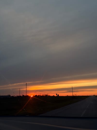 The image captures a vibrant sunset or sunrise over a semi-rural landscape, viewed from the interior of a moving vehicle, likely in Alexandrovka, Russia.
The sky dominates the upper two-thirds of the frame, showcasing a spectacular display of warm colors. Layers of clouds are illuminated in brilliant shades of orange, deep yellow, and red near the horizon, gradually transitioning to softer grays and blues higher up. The sun, a bright, intense orb, is visible in the lower right quadrant, partially obscured by the distant landscape, casting strong rays and creating noticeable lens flare effects.
Along the horizon, a dark silhouette defines the land, featuring sparse trees, bushes, and various man-made structures. Prominent among these are utility poles and at least one tall communication or cellular tower, standing starkly against the glowing sky. A straight road, likely an asphalt highway, occupies the right side of the image, with a white dashed line marking the lane, extending into the distance. The dark, curved edge of a vehicle's interior is visible on the far right, framing the scene.
The time of day is clearly sunset or sunrise, characterized by the low position of the sun and the dramatic, colorful sky. The weather appears clear with scattered clouds. There is no visible text in the image. The scene evokes a sense of tranquility and the natural beauty of the day's beginning or end.