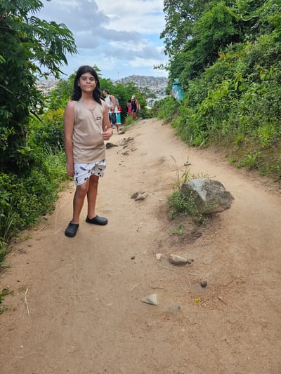 The image displays a hiking trail on a vegetated hillside in Arraial do Cabo, Brazil, presenting a view of the urban area and distant water under an overcast sky. In the foreground, slightly left of center, an individual identified as a child, approximately 8-12 years old, stands facing the camera. This child has long dark hair and is dressed in a light beige sleeveless top, white shorts adorned with dark patterns, and black clogs. Their right hand is raised, performing a thumbs-up gesture. Further along the winding, reddish-brown dirt path, several adults are visible walking away from the viewer. Among them, a male figure in a light shirt carries a backpack, and a female figure in dark attire carries a bag. The path is uneven, interspersed with small rocks and patches of green vegetation. A prominent grey boulder, surrounded by grass and small plants, rests on the right side of the path in the mid-ground. Dense green foliage and trees line both edges of the path. A teal-colored cylindrical refuse bin is partially visible, embedded within the vegetation on the left slope. The background features a cityscape with numerous buildings clustered on hillsides, extending towards what appears to be a body of water, beneath a sky dominated by white and grey clouds.