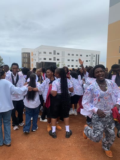A group of young adults, likely graduates, are celebrating outdoors in front of a modern, multi-story building. Many are wearing white shirts covered in handwritten messages and signatures. The ground is covered in reddish-brown dirt. The sky is overcast. Some individuals are interacting with each other, with some shaking hands or embracing. One person in the foreground, wearing a black beret and a white shirt with many signatures, is looking at the camera and smiling broadly, holding a red scarf. Another person in the foreground is wearing patterned trousers and orange boots. The building in the background appears to be an educational institution. This scene is located in South Ropp, Nigeria.