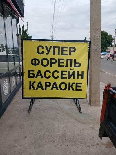 In Kara-Balta, Kyrgyzstan, a bright yellow banner with black Cyrillic text is prominently displayed on a metal easel in front of a building with large glass windows. The banner reads "СУПЕР ФОРЕЛЬ БАССЕЙН КАРАОКЕ," which translates to "SUPER TROUT POOL KARAOKE." The text is large and bold, making it easily readable from a distance. The building appears to be a commercial establishment, with what looks like food-related graphics visible on the glass windows, including a pizza slice, a burger, and an umbrella.

To the right of the banner, a concrete utility pole stands tall, showing signs of wear and tear. Beyond the pole, a paved road is visible, with a few parked cars and some pedestrians walking. In the background, trees and buildings complete the urban landscape under a cloudy sky. The overall scene suggests a business advertising its services in a public area of the city.