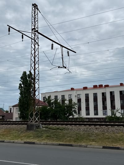 The image depicts a cloudy day in Derbent, Russia, with a focus on railway infrastructure and urban development. In the foreground, a paved road with a curb runs horizontally, bordered by a grassy verge. Behind the verge, railroad tracks are visible, with ballast stones and adjacent vegetation. A tall, metal lattice power pole stands prominently near the tracks, supporting overhead electrical wires for a railway system. The pole is weathered and shows signs of rust.
In the background, a modern, multi-story apartment building with a red-tiled roof and white facade dominates the scene. The building features numerous windows with dark frames and a row of distinctive chimney-like structures along the roofline. To the left of the apartment building, a tall, leafy tree is situated, partially obscuring a smaller, darker-roofed residential building. The overall environment suggests an area where urban expansion meets transportation networks, under a overcast sky. No people or animals are visible in the frame.