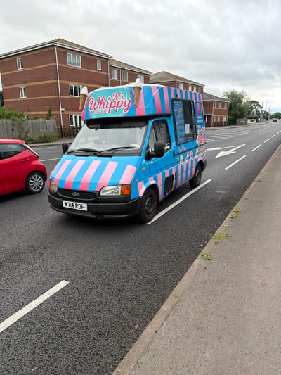 In a suburban area of Gloucester, United Kingdom, an ice cream van with blue and pink stripes is parked on the side of a road. The van is branded with "Mr Whippy Gloucester" and features images of ice cream cones. A red car is visible to the left of the van, and residential buildings can be seen in the background. The sky is overcast, and the road appears to be wet, suggesting recent rain. The scene depicts a typical day with the ice cream van operating in a residential neighborhood.