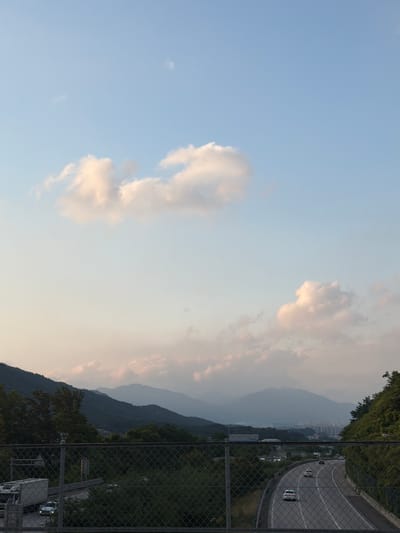 The image depicts a panoramic view of a highway in Pocheon-si, South Korea, during a twilight hour. The sky is a gradient of soft blue and pale pink, with scattered fluffy clouds illuminated by the setting sun. In the distance, a range of mist-covered mountains forms a hazy silhouette against the sky. The highway curves gently through the landscape, with several cars visible on its lanes. The foreground features a chain-link fence and lush green trees, with a white truck parked on the left side of the road. The overall atmosphere is serene and tranquil, characterized by the interplay of natural beauty and human infrastructure.