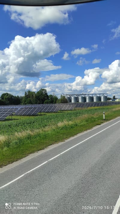 The image depicts a rural landscape on a bright, partly cloudy day. In the foreground, a gray asphalt road with a white dividing line runs diagonally from the bottom left to the top right. Adjacent to the road is a grassy verge with dry, tall grasses on the left. Further left, a large field of solar panels stretches horizontally across the middle ground. Behind the solar panels, a line of green trees forms a backdrop. In the distance, several tall, cylindrical silver silos are visible, along with some smaller buildings. The sky is a vibrant blue, dotted with fluffy white clouds. The timestamp "2026/7/10 15:17" and "REDMI NOTE 9 PRO AI QUAD CAMERA" are present in the bottom right and bottom left corners, respectively. This scene is located in the vicinity of Imavere, Estonia.