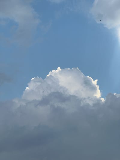 An outdoor, eye-level, full shot captures a partly cloudy blue sky on a bright day. In the distance, a small bird can be seen flying. The bottom half of the image is dominated by dark, heavy clouds, while the top half features a bright, luminous white cloud formation. The sun is obscured by clouds, but its rays are visible as a bright light emanating from the top right corner of the frame. The image does not depict any elements specific to the city of Straubing, Germany.