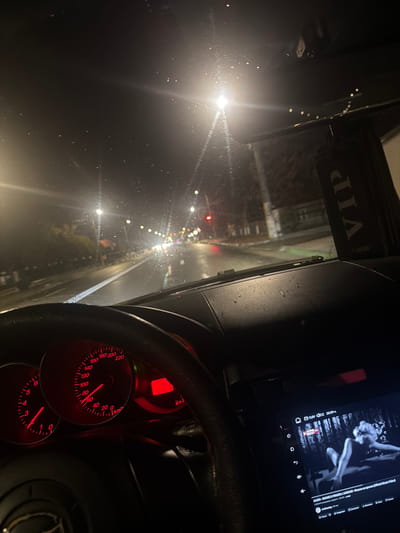 The image depicts the interior of a car, presumably at night, as seen from the driver's perspective. The scene includes the car's dashboard with glowing red gauges, a steering wheel, and a digital display screen showing a music video playing. In the windshield's view, we can see a wet road with blurred streetlights, indicating the car is likely in motion. A "VIP CARIBI" sticker is affixed to the top of the windshield. The OCR shows the car is going 65 km/h, the date and time is 9/22 20:59, and is playing a song by ALISIA. There is no evidence to indicate this is the city of Tuzla, Romania.