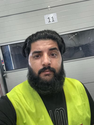 A man with dark hair and a full beard, wearing a neon yellow vest over a black t-shirt with white lettering, is pictured from the chest up. He is standing in front of a large, ribbed, grey overhead door, with a horizontal window panel above. A white rectangular sign with the number "11" in black digits is affixed to the door above and to the left of the man's head. The scene appears to be an industrial or warehouse environment.
