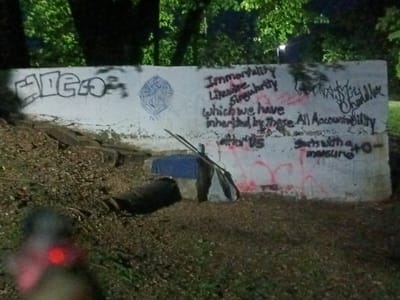 An outdoor, low-light photograph taken in Tahlequah, United States of America, depicts a large concrete retaining wall covered in graffiti. The time of day appears to be dusk or night, with visible light sources in the distant background suggesting artificial lighting. Lush green foliage and tree trunks are visible above the wall and in the background, contrasting with the dirt and grass-covered ground in the foreground and at the base of the wall.

Leaning against the wall at ground level is a rudimentary, makeshift shelter constructed from a blue tarp and white fabric, supported by several sticks or poles. A dark, possibly rolled-up object lies on the ground near this shelter. In the immediate blurry foreground, a red-glowing object is partially visible.

The concrete wall is extensively graffitied. On the far left, in large block letters, the word "AIGO" is spray-painted in black. Towards the center, there is a complex, circular design. To the right of this design, a multi-line message in black spray paint reads:
"Immortality"
"Likewise"
"Singularity"
"which we have"
"inherited by those. After US."
Further to the right, another message, also in black spray paint, states:
"All Accountability"
"starts with a +0-"
"measure"
Above this text, additional illegible tag-style graffiti is present. There are also several red spray paint marks and splatters visible on the lower portion of the wall.