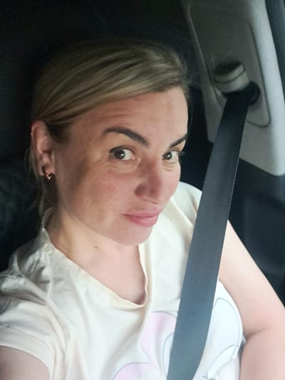 A woman is captured in a close-up, horizontal shot inside what appears to be a car, possibly in Mayak, Russia. She has blonde hair, fair skin with some visible freckles, and dark, defined eyebrows framing her dark eyes. Her gaze is directed straight at the camera, and her expression is neutral. She wears a light-colored t-shirt, possibly white or cream, featuring faint light pink and grey graphic outlines on the right side of her chest. A black seatbelt diagonally crosses her torso and shoulder, leading towards a grey car interior panel with a visible seatbelt anchor point in the upper left corner of the frame. The woman appears to be reclined or lying down, with her head turned to face the camera. A small earring is visible in her left ear. The lighting is soft and diffused, characteristic of an interior setting. There is no visible text in the image.