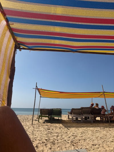 The image presents a beach scene in Bhagatwadi, India, viewed from under a striped fabric canopy displaying blue, red, and yellow horizontal bands. A human leg is visible in the bottom-left foreground. The sandy midground features another, smaller striped canopy, supported by wooden poles, exhibiting red, yellow, orange, green, and blue horizontal stripes. Beneath this secondary canopy, several wooden lounge chairs are arranged. Three individuals are partially visible: one wearing a light-colored bucket hat, another (possibly a child) in a light-colored hat and patterned top, and a third person seated to the right. All individuals appear to be resting. The background consists of a calm, blue-green ocean extending to the horizon under a clear, light blue sky.