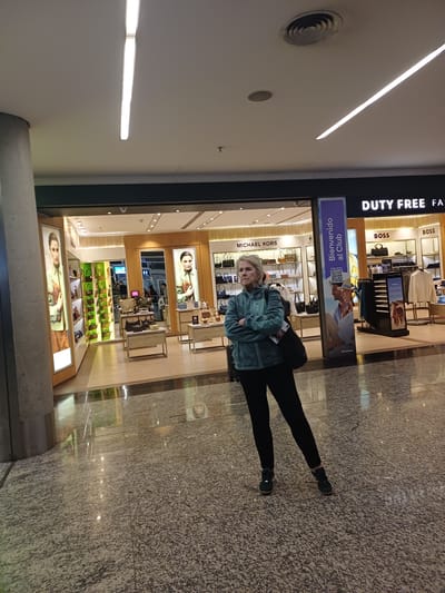 A medium shot shows an elderly Caucasian woman standing with her arms crossed in an airport terminal in Aeropuerto Internacional Ezeiza, Argentina. She is wearing a teal fleece jacket over black leggings and dark sneakers. A black bag is slung over her right shoulder. She is looking off to her left with a neutral expression. Behind her, a "DUTY FREE" shop is visible, with displays of handbags and clothing. A sign for "MICHAEL KORS" is also visible. The floor is tiled with a speckled pattern. The lighting is bright, with long fluorescent lights visible on the ceiling. The overall environment appears to be a modern airport terminal with shops and signage.