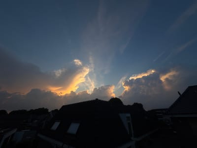 This image depicts a twilight sky over the city of Putten, Netherlands. The sky is a deep blue, transitioning to lighter hues near the horizon, where dramatic clouds are illuminated by the setting sun. Streaks of light, known as crepuscular rays, emanate from behind the clouds. In the foreground, the dark silhouettes of rooftops are visible, indicating a residential area. A bird is perched on the roof of one of the houses on the right side of the image. The overall scene suggests a peaceful evening in a Dutch city.