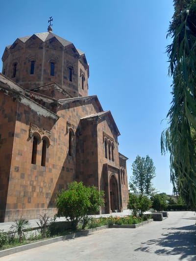 The image shows a side view of a large, ornate, ancient stone church with a dome topped by a cross. The church is constructed of reddish-brown stones and features intricate carvings around its arched windows and entrance. The sky is a clear, bright blue. In the foreground, there is a paved walkway with a landscaped garden bed containing small trees, bushes, and flowering plants. To the right, the branches of a weeping willow tree hang into the frame, casting dappled shadows on the walkway. The scene appears to be located in Artashat, Armenia, possibly at the Mother See of Holy Etchmiadzin complex, given the architectural style and religious iconography. No people are visible in the image.