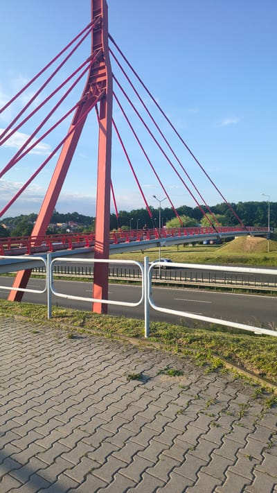 The image captures a view of a pedestrian and bicycle cable-stayed bridge, painted a vibrant red, against a clear blue sky. The bridge features a prominent red pylon, from which numerous red cables extend to support the bridge deck. A white metal railing runs along the edge of the sidewalk in the foreground. Beyond the railing, a multi-lane road is visible, with a silver car driving on it. Green grass and trees form the background, with a distant view of buildings and a church spire on a hill, suggesting the location is near a town or village. The foreground shows a pathway paved with interlocking concrete blocks. The lighting suggests it is daytime, likely late afternoon due to the long shadows. This scene is located in Góra Świętej Anny, Poland.