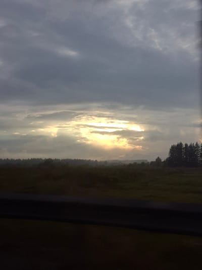 The image depicts a dramatic sunset scene over a rural landscape, likely in or near Anikino, Russia. The sky is filled with heavy, dark clouds, but a break in the cloud cover allows shafts of bright, golden sunlight to stream through, illuminating the horizon. The light creates a halo effect around the illuminated areas, casting a warm glow.

Below the sky, a dense line of dark trees forms a treeline, suggesting a forest or wooded area. Beyond the treeline, the landscape appears to be a field or open grassland, with a darker, shadowed foreground. A dark, horizontal object, possibly a guardrail or a fence, is visible in the lower portion of the image, suggesting a road or a viewpoint overlooking the scene. The overall atmosphere is one of dramatic natural beauty, with the interplay of light and shadow creating a sense of depth and mood.