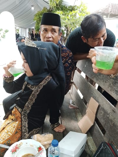 The image captures an outdoor social gathering in Juwana, Indonesia, beneath a white fabric canopy. In the foreground, a young individual, likely a child, sits on a rustic wooden bench. They are dressed in a black traditional garment with ornate gold embroidery and a black hijab, embellished with a silver tiara-like accessory, while drinking a light green liquid from a clear plastic cup. Directly behind this individual is an older man wearing a black peci and a brown batik-patterned shirt, looking towards the left side of the frame with a focused expression. To his right, another man, younger, leans forward onto the wooden bench back, holding a clear plastic cup containing the same green liquid and observing it with a slight smile.
On the bench to the left of the young person, a white plate with a floral design contains a small amount of food and a spoon. Adjacent to it are a bottled water with a blue cap, a clear plastic bag holding light-colored snacks, and a white cardboard box. A black quilted bag is visible on the far right, partially obscured by the bench slats. A piece of bent cardboard and a small plastic bag are on the ground beneath the bench. The background is blurred, showing additional attendees, green foliage, and distant roof tiles, suggesting a residential or communal outdoor area.
