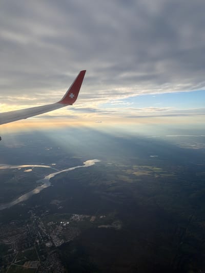 The image shows an aerial view from an airplane window, looking down at a landscape during what appears to be either sunrise or sunset. A portion of the airplane's wing, painted white with a red tip and featuring a white star emblem, is visible in the upper left quadrant of the image.
The landscape below is a mix of urban and natural environments. A winding river, reflecting the light, cuts through the terrain. On the left side of the river, there is a developed urban area with buildings and roads, consistent with a town or city. On the right side, the land is covered by a dense, dark green forest, suggesting a rural or less developed area.
Sunlight is breaking through the clouds in distinct rays, illuminating parts of the landscape and creating a dramatic effect. The sky is filled with clouds of varying shades of gray and white, with a strip of blue sky visible near the horizon.
The location is identified as the city of Dubrovo (Uleiminskoe S/P), Russia. This suggests the aerial view captures this specific region of Russia. There are no people or specific actions depicted in the image, only the static scene of the landscape and the airplane's wing against the sky. The focus is on the geographical features and the atmospheric conditions.