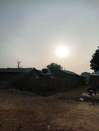 The image displays an outdoor scene in Nata-Ala, Nigeria, characterized by a bright, low-angle sun positioned slightly right of the frame's vertical center against a hazy, pale grey to yellow sky. The foreground comprises an expanse of reddish-brown, dry earth. In the mid-ground, multiple low-rise structures, appearing to be constructed from earthen materials with dark, possibly corrugated roofs, are visible and heavily shadowed. Two distinct buildings are on the left, with additional structures receding into the background on the right. A dark, leafless tree silhouette is present on the upper left, while dense green foliage from another tree occupies the upper right. On the lower right, a black circular object, resembling a tire, rests on a raised concrete base, next to which a horizontal metal rod extends. Scattered light-colored debris is visible on the ground near this object. No individuals are present.