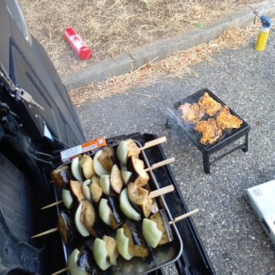 This image shows an outdoor scene where food is being prepared on a grill. In the foreground, a metal tray holds skewers of what appear to be grilled eggplant and onion, interspersed with some pieces of potato. To the right, a small black grill with legs is set up on the asphalt ground, with several pieces of grilled chicken on its rack. Smoke rises from the grill, indicating that the chicken is actively cooking.
In the background, dry, straw-like grass and a concrete curb are visible, suggesting a somewhat rustic or informal outdoor setting. A red canister, possibly a fire extinguisher, lies on the ground next to the grass. A yellow butane gas canister with a torch attachment is also present, positioned near the curb. The lighting suggests it is daytime, likely late afternoon or early evening, with natural light illuminating the scene.
To the left of the image, part of a dark-colored vehicle, possibly a car trunk or storage compartment, is visible. Keys are hanging from a latch on this compartment. A pack of cigarettes, with Spanish text on it that reads "El humo del tabaco contiene más de 70 sustancias cancerígenas" (Tobacco smoke contains more than 70 carcinogenic substances), is also placed near the food tray. The overall impression is a casual outdoor cooking event, perhaps a barbecue or picnic.