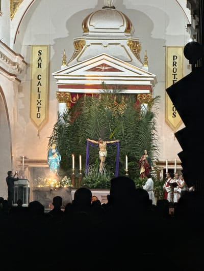 An indoor photograph depicts a religious scene, likely a procession, within a church in Timaná, Colombia. The altar, adorned with ornate gold and white decorations and signage reading "SAN CALIXTO" and "PROTEOS", displays religious figures. A statue of Jesus on the cross is prominently featured, surrounded by palm fronds and lit candles. A statue of Mary in blue is also visible. Several people stand in the background, obscured by black silhouettes and the darkness of the scene. A few individuals appear to be holding phones, possibly recording the event. The atmosphere is dim and reverent.