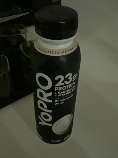 A black Yopro protein drink bottle lies horizontally. It's labeled "23g Protein" with added magnesium and vitamin B6. The flavor, "Stracciatella Geschmack," is printed near the black cap. The label indicates "0% Zuckerzusatz" (no added sugar) and "0% Fett" (0% fat).