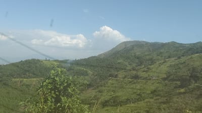 Green mountains stretch under a blue sky dotted with fluffy white clouds. The slopes are covered in lush vegetation, with varying shades of green indicating different types of trees and plants. A dirt road winds its way through the hills. In the foreground, some grass and shrubs are visible. Power lines can be seen in the top left corner of the image.