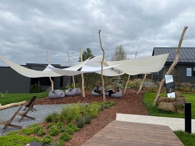 The image depicts an outdoor seating area with a contemporary design, situated in Yakunino, Russia. A large, white, fabric canopy, supported by rustic wooden poles, provides shade over a seating arrangement of gray beanbag chairs. Two individuals are seated in the beanbag chairs, appearing relaxed and engaged in conversation. In the foreground, a wooden deck extends into the scene, adjacent to a gravel area with two wooden lounge chairs. A well-maintained garden with a variety of plants and flowers borders the seating area. In the background, a modern building with dark siding and a gray roof is visible. The sky is overcast, suggesting a cloudy day. The overall atmosphere is serene and inviting, designed for outdoor leisure.