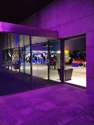 A building illuminated in vibrant purple light features a glass entrance. People are visible through the glass doors. One doorway displays "PLAZA" signage internally. A potted plant sits near the entrance. The purple hue dominates the façade, sidewalk, and even reflects within the glass, creating a monochromatic effect.