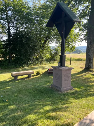 This image depicts a memorial cross situated in a grassy area in Momberg, Germany. The cross, made of dark wood, stands on a stone pedestal. Inscriptions are visible on the pedestal in German. To the left of the cross, a wooden bench rests on two stone supports. The environment is characterized by lush green grass, trees, and a sunlit landscape in the background with a road and a hay bale. There are no people or animals present in the image.