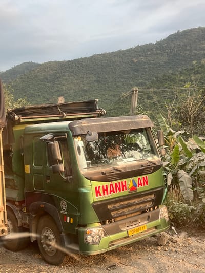 A green "KHANH AN" truck with license plate 79H-078.11 is parked on a dirt or gravel road. The truck's front end is visible, showing the grill, headlights, and front tires. The road is adjacent to a lush green mountainside and foliage. The photo was taken in Aê Thi, Vietnam. A telephone pole with wires is also in the background.