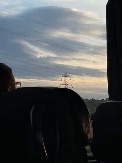 The image shows a view from inside a vehicle, likely a car or bus, looking out towards the sky. Overhead power lines and a utility tower are visible against a backdrop of cloudy skies, possibly indicating an overcast day. In the foreground are parts of the vehicle's interior, including a headrest and the top of a person's head, partially visible. The location is likely in the Surcin Urban Municipality, Serbia, due to the presence of the utility tower and overhead power lines, which are common infrastructure elements.