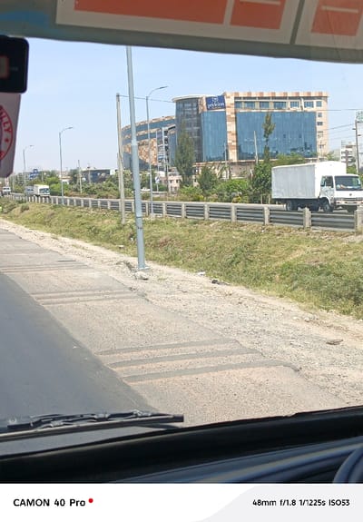 Captured from a vehicle, the scene features an asphalt road, transitioning into a rougher, patched surface. A grassy median with a metal guardrail separates the road from a distant building with a modern, glass-clad facade. A white truck travels along the road. Lampposts punctuate the landscape. The windshield's edge is visible at the bottom, providing a driver's perspective on a bright, sunny day.