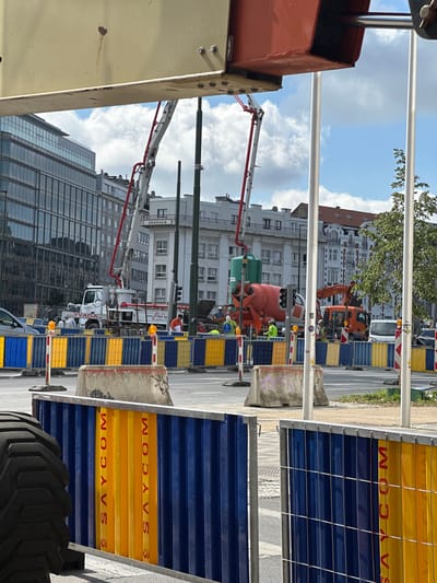 The image depicts a street scene in Molenbeek-Saint-Jean - Sint-Jans-Molenbeek, Belgium, characterized by ongoing construction work. A large concrete mixer truck with a pump attachment is positioned in the center of the frame, with its boom extended. Adjacent to it, another construction vehicle, possibly an excavator, is also present. Several workers in high-visibility vests are visible, engaged in construction activities. The area is cordoned off with temporary barriers, including concrete blocks and fencing with blue and yellow panels bearing the text "SAYCOM". In the background, modern and older architectural styles of buildings are visible, indicating an urban environment. The sky is partly cloudy. The overall scene conveys a sense of active infrastructure development.