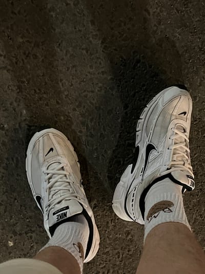 Two white Nike "Initiator" sneakers with black accents and white socks are visible, standing on a dark, textured asphalt surface. The left shoe is closer to the viewer and is angled slightly outwards. The right shoe is further away and angled more inwards, with the wearer's right leg also visible. The socks have a graphic design on them that appears to be a cartoonish brown object with red lettering underneath. The background is a dark, out-of-focus asphalt surface. The image appears to be taken at ground level. The location is Strumivka, Ukraine.