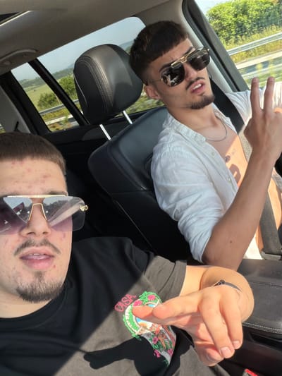 Two young adult males are depicted inside a vehicle on a bright, sunny day.
The male in the foreground, positioned in the driver's or front passenger seat, faces the camera. He has short, dark hair with a fringe, a trimmed goatee, and a mustache. His forehead and cheeks display visible acne. He wears large, gold-framed aviator-style sunglasses with dark, reflective purple-tinted lenses that obscure his eyes. His mouth is slightly open. He is dressed in a black short-sleeved t-shirt with a colorful graphic design on the right side of the chest that includes the word "CASA" in pink. His left hand, positioned in the lower right quadrant of the image, is extended forward, forming a finger gun gesture, and he wears a dark-banded wristwatch.
The second male is seated in the backseat on the right side of the frame, turned slightly towards the camera. He also has short, dark hair with bangs and a trimmed beard and mustache. He wears gold-framed aviator sunglasses with dark, highly reflective lenses. His mouth is slightly open, revealing his teeth. He is wearing a white, unbuttoned long-sleeved shirt, revealing a silver chain necklace and a tattoo of Arabic script on the right side of his chest. His right hand is raised, forming a peace sign (V-gesture). A black seatbelt crosses his chest, with the "PRE-SAFE" label visible on the buckle.
The vehicle interior features black leatherette seating, with a prominent headrest visible behind the front passenger. Through the car windows, a clear blue sky and a green landscape are visible, including trees, fields, and distant hills. A road with a guardrail runs alongside the vehicle. The scene is illuminated by direct sunlight, casting strong shadows within the car.
The scene is located in Mlada Gvardia, Bulgaria.