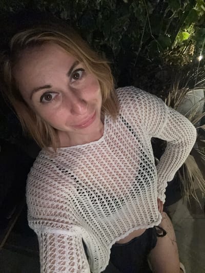 A fair-skinned woman with blonde hair is lying down, facing the camera. She is wearing a white, long-sleeved, open-knit top over a black top. Her expression is neutral. The background includes dark foliage and what appears to be a palm frond. Part of a leg is visible in the upper right corner, with dark-colored clothing underneath. The setting seems to be outdoors, possibly in Dobra Voda, Montenegro, given the user's prompt.
