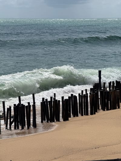 The image captures a view of the sea and beach at Ban Tha Chat Chai, Thailand. In the foreground, a sandy beach is lined with a series of weathered, dark wooden posts, partially submerged in shallow water. These posts appear to be remnants of a pier or some form of coastal defense. Beyond the posts, a large wave is crashing, with white foam and spray filling the area between the posts and the open sea. The ocean water is a greenish-blue color and appears choppy. In the distance, the sea meets a hazy sky, with sunlight glinting off the water's surface. The overall scene suggests a rugged and dynamic coastal environment.