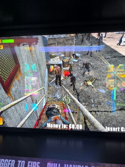 The image displays a brightly lit screen of a zombie-themed arcade video game, likely captured from an angled perspective looking down onto the action. The game depicts a grim, outdoor urban environment, possibly a shantytown or industrial zone, characterized by corrugated metal fences/walls and debris.

Numerous figures, appearing as zombies, are scattered across the scene. In the immediate foreground, a defeated zombie lies face down on a reddish metal platform or walkway, with its body distorted. Further back, several other zombies are standing or moving, some appearing to be injured or slumped. One zombie stands prominently near a stack of what looks like discarded items, while another is partially obscured by a wheelbarrow. More zombie figures are visible in the distance.

The activity captured is mid-gameplay. The player's perspective is from an elevated position, overlooking the chaotic scene. Game interface elements are visible: a red health bar with a green segment and the number "23" are in the top-left corner. Various glowing, colorful graphical elements, possibly targeting reticles or power-ups, are scattered throughout the screen.

Visible text on the game screen includes "Money In: £0.00" and the partially cropped "Insert Co" (likely "Insert Coin") on the bottom right. Below the screen, the arcade machine itself shows text: "TRIGGER TO FIRE PULL HANDLE." The location context provided indicates this arcade machine is in Agios Dimitrios, Greece.