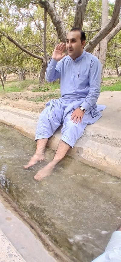 A man, wearing a light blue shalwar kameez and a wristwatch, sits with his feet submerged in a shallow water channel. He is facing to the right, with his right hand raised to his ear as if listening. The man is positioned on the edge of a concrete structure overlooking the water. The environment appears to be outdoors, with trees and greenery in the background, suggesting a rural or park-like setting. The lighting indicates daytime. The scene is likely located in Kan Mehtarzai, Pakistan.