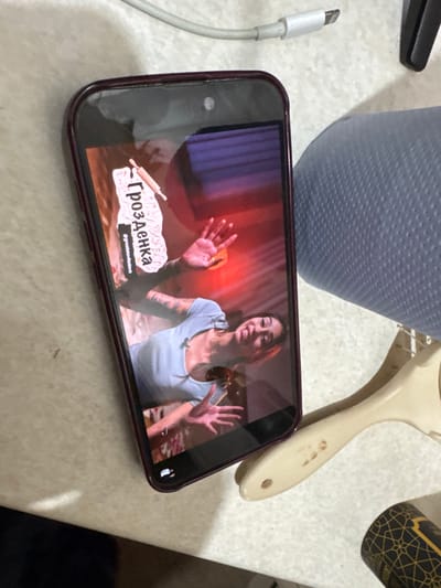 A top-down photographic view captures an indoor scene in Fowlis Easter, United Kingdom. A dark-cased smartphone, likely an iPhone, is positioned vertically on a light-colored, possibly beige, flat surface. The phone's screen displays a video featuring a young woman with dark hair and visible arm tattoos, wearing a light blue top with a small microphone clipped to the collar. She is smiling and has both hands raised with palms open, against a background illuminated with red and purple lighting. In the upper left of the screen, a logo with Cyrillic text "ГроЗденка" (GroZdenka) and a rolling pin icon is visible, along with the smaller text "@grozdankadudova". A white USB-A to Lightning cable is present above the phone, with its connector near the device. To the right of the smartphone, a textured blue cylindrical object, possibly a rolled towel or cushion, is partially visible. Below this, a cream-colored object resembling a hairbrush handle with minor dark staining is present. A dark cylindrical object featuring a gold geometric pattern is partially visible in the bottom right corner of the frame.