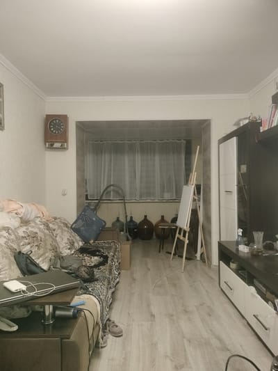 This image captures the interior of a residential living space in Sheptytskyi, Ukraine. The room features light-colored walls, some with visible wallpaper texture, and a white ceiling with crown molding. The floor is covered with light wood-laminate planks.
To the left, a large sofa with a beige floral pattern is partially covered by a dark, patterned throw blanket. Piles of light-colored clothing are present on the sofa. On a small, dark side table or armrest extension, a closed dark laptop is visible, topped with a white power bank and white cables. Below this, a dark hairdryer is plugged in, alongside a blue comb-like object. A pair of light-colored sneakers, likely worn by a person seated or reclining on the sofa, are visible on the floor next to the sofa. A vintage-style rectangular wooden wall clock with Roman numerals hangs on the wall above the sofa, and a light switch or outlet cover is visible below it.
The center of the room opens into a recessed area, possibly a converted balcony or alcove, defined by a wide, framed entryway. This area features a window at the back, covered by sheer white curtains. Inside this recess, from left to right, are two large, dark brown glass demijohns or carboys of varying shapes (one spherical, one with a narrower neck), a small, dark, round table with thin legs, and a wooden artist's easel holding a blank white canvas. A dark, flexible vacuum cleaner hose is coiled and leaning against the wall near the left demijohn. A cardboard box is on the floor near the right demijohn.
The right side of the room contains dark brown furniture units. A tall wardrobe or cabinet with a white door section is on the left, and a lower console or TV stand with two white drawers/cabinets is on the right. On the surface of this console, a clear spray bottle, a small clear glass pitcher containing a dark liquid, and other small items are visible. A dark bookshelf with books and decorative objects is mounted above these units at the top right of the frame. No individuals are clearly depicted, though the presence of sneakers suggests a person is occupying the sofa, largely obscured by other items.