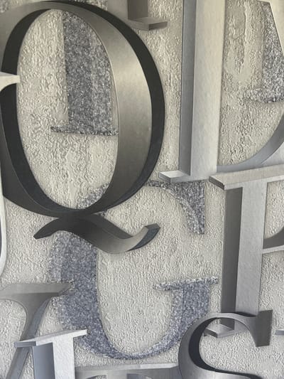 The image displays a close-up view of a textured wallpaper or fabric with a modern, abstract design. The design features bold, three-dimensional geometric shapes and stylized letters in shades of grey and silver against a textured off-white background. The texture of the background appears to be a raised, uneven pattern, possibly mimicking stucco or a rough fabric weave.
Some of the shapes resemble stylized letters, such as a large, curved "S" or "2" shape in the foreground, rendered in dark grey with a metallic sheen. Other elements include sharp, angular forms in lighter grey, some with a textured, glittery inlay of small silver particles. These elements are arranged in an overlapping and layered composition, creating a sense of depth. There are no people, animals, or discernible objects other than the abstract design elements themselves.
The lighting appears to be even and diffused, suggesting an indoor setting, perhaps a wall in a room. There are no indications of time of day, weather, or specific location cues within the image itself, other than the context provided that it relates to Geamăna, Moldova, which is not visually evident in the image. No text is legible. The overall impression is one of a contemporary and sophisticated decorative surface.