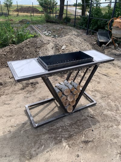 The image depicts an outdoor scene in what appears to be a construction or renovation area. A custom-made metal barbecue grill with a white tiled side is the central focus. The grill is elevated on a dark metal frame that has a triangular, X-shaped support structure. This support structure also serves as a wood rack, with several logs of firewood stacked within it.
The surrounding environment is characterized by loose dirt and gravel, indicative of ground disturbance. Piles of soil are visible in the background, along with some construction materials like wooden planks. A black metal fence runs along the perimeter of the area, with trees and green foliage beyond it. In the far background, a concrete mixer can be seen, suggesting ongoing construction work. The sky is overcast, hinting at a possibly humid or pre-rain weather condition. The scene is devoid of any people. The overall impression is one of a functional, yet unfinished, outdoor space.